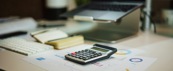 A calculator rests on printed financial charts with colorful graphs, placed on a modern office desk next to a notebook, keyboard, and open laptop on a stand. The workspace suggests a financial planning or debt management setting, aligning with services like Credit Associates.