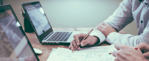 Two people review handwritten notes and financial sketches at a desk with open laptops, one pointing with a pencil while discussing the content. The setting suggests a collaborative session focused on debt relief or financial strategy, aligning with services like TurboDebt.