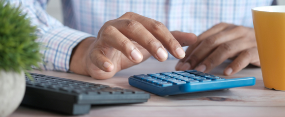 Close-up of a person in a plaid shirt using a blue calculator at a desk, with a keyboard, yellow coffee mug, and a small potted plant nearby. The image reflects a personal finance or budgeting moment, relevant to services like Pacific Debt Relief.