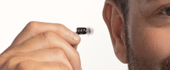 Close-up of a man holding an Eargo hearing aid near his ear, showing the small, in-canal design with soft, flexible tips. The device is black and discreet, highlighting Eargo's nearly invisible hearing technology.