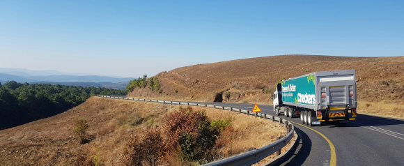A semi-truck drives along a winding rural highway bordered by guardrails, dry hills, and scattered trees under a clear blue sky. This image illustrates fleet movement and logistics operations, relevant to Verizon Connect’s focus on fleet management and route efficiency.