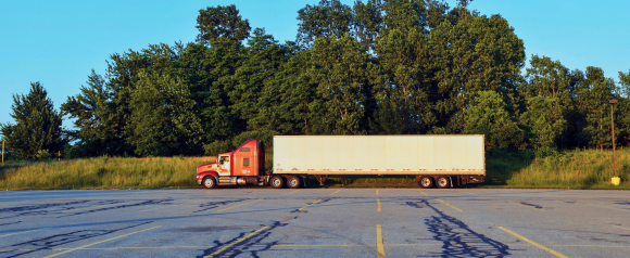 A red semi-truck with a white trailer is parked in an empty lot near a line of tall green trees during golden hour. This image highlights a moment of rest or staging in fleet operations, aligning with Azuga’s focus on fleet tracking and location monitoring.