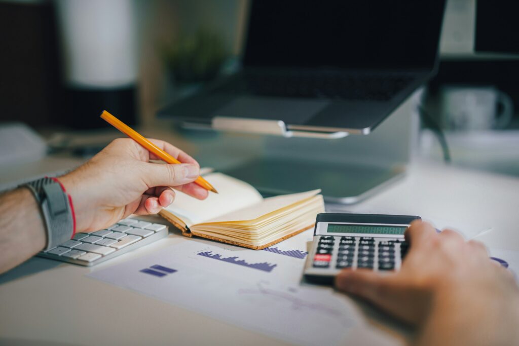 Person using a calculator while taking notes in an open notebook, with printed financial graphs on the desk and a laptop in the background. The scene conveys financial organization, relevant to understanding how does debt consolidation work.