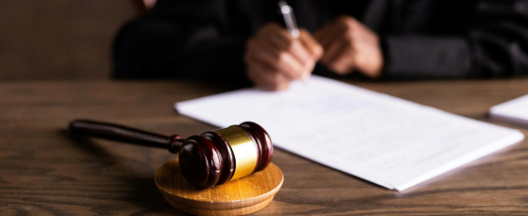 A wooden judge's gavel rests on a block in the foreground while a person in dark robes signs legal documents in the background. The image conveys a legal setting, reinforcing themes of justice and courtroom action, aligning with Marcus and Zelman as class action lawyers.