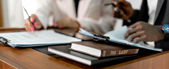 Two professionals review documents together at a wooden desk, with a law book labeled “THE LAW” and binders in the foreground. The image evokes a collaborative legal setting, reinforcing the expertise of Pollock Cohen LLP as class action lawyers.