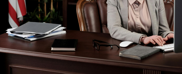A professional in a light gray suit types at a desktop computer in a law office, with legal documents, notebooks, and glasses on a polished wooden desk. The organized workspace conveys focus and diligence, aligning with the mission of The Consumer Lawyers.
