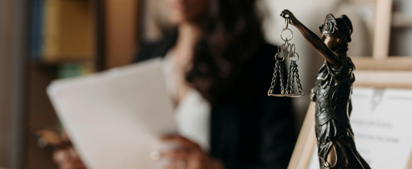 A close-up of a bronze Lady Justice statue holding balanced scales, with a blurred figure reviewing legal documents in the background. The scene symbolizes fairness and legal integrity, supporting the reputation of Siri and Glimstad as class action lawyers.