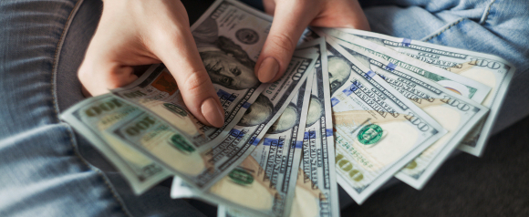 A person fans out several $100 bills while sitting in blue jeans, highlighting a sense of financial gain or careful money management. The image represents the financial empowerment and support services offered by Elevate 1 Financial.