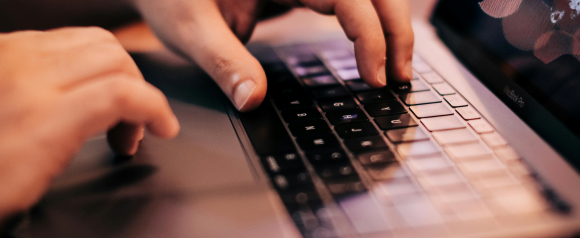 Close-up of hands typing on a MacBook Pro keyboard, with a blurred screen showing abstract shapes. The image suggests digital work and backend operations related to consent management platforms like iubenda.