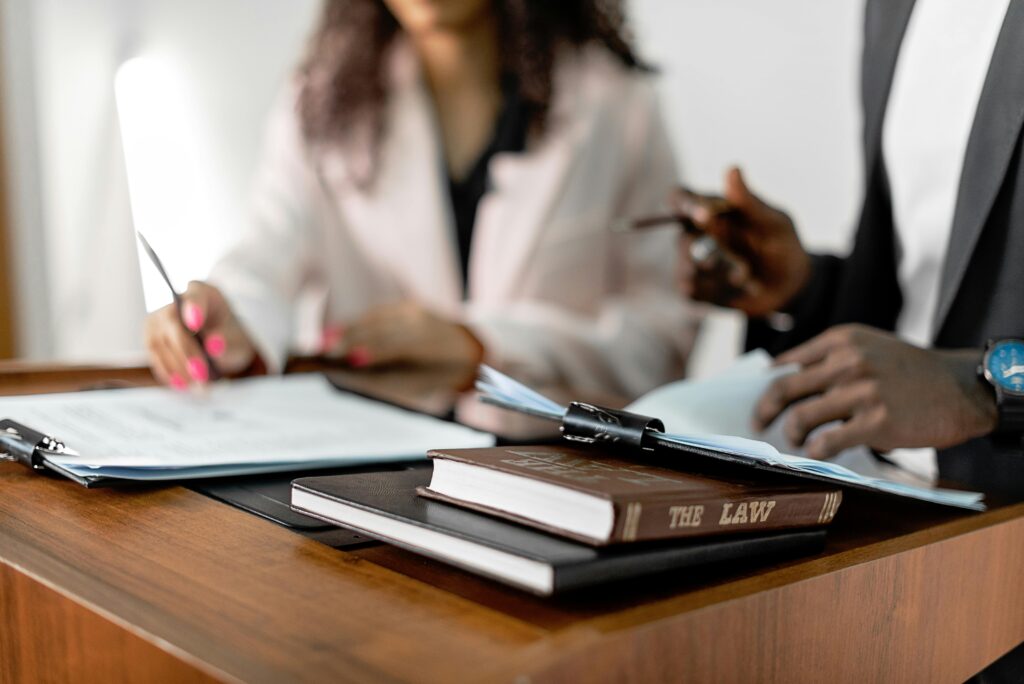Close-up of two professionals reviewing documents at a desk, with a law book titled "The Law" prominently placed in the foreground. The image conveys a legal setting, suggesting class action lawyers working on case files or legal strategy.