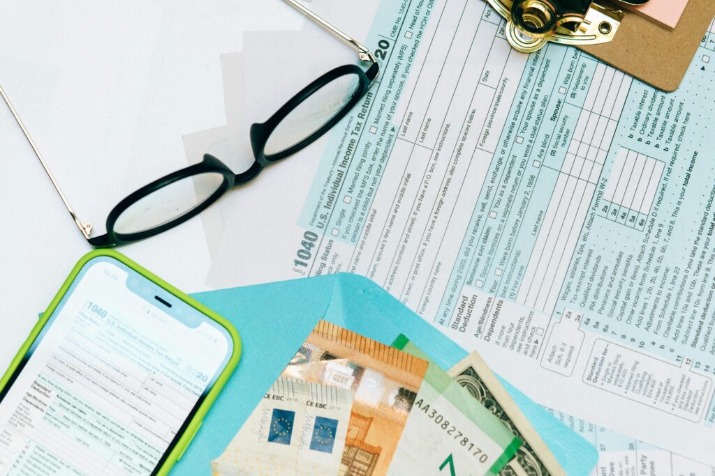 A flat lay of U.S. 1040 tax forms from 2020 alongside reading glasses, a smartphone displaying the same form, and various international currencies in a blue folder. The scene suggests financial preparation related to tax topic 152 Refund Information.