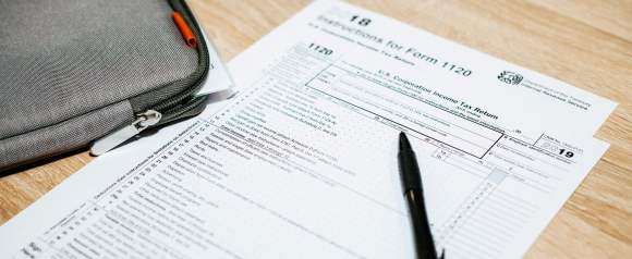 A 2019 U.S. Corporation Income Tax Return Form 1120 rests on a wooden desk beside a gray zippered pouch and a black pen. This setup suggests tax filing or preparation, a key service area for Larson Tax Relief.