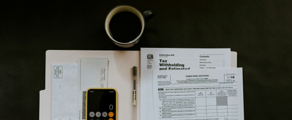 Overhead view of tax preparation materials including IRS tax forms, a smartphone calculator, a Muji pen, a coffee mug, and a folder, representing a typical setup for organizing finances or working with a tax relief service like TaxRise. Prominently visible forms include “Tax Withholding and Estimated” and “Capital Gains and Losses.