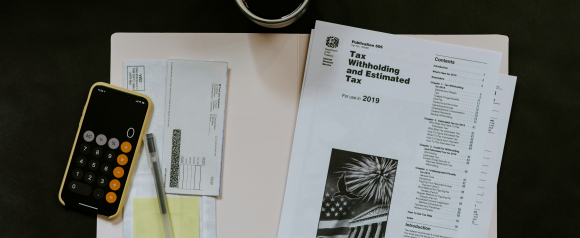 A smartphone with a calculator app open, tax documents, a pen, and an IRS publication titled “Tax Withholding and Estimated Tax” for 2019 are arranged on a desk. The setup reflects personal tax preparation or financial review, relevant to services like Tax Hardship Center.