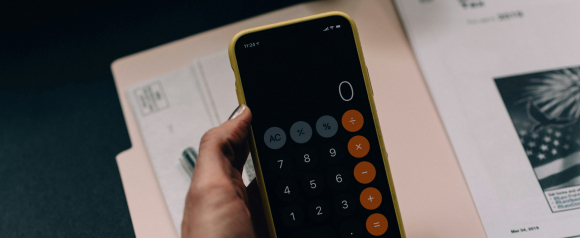 Close-up of a hand holding a smartphone with a calculator app open, displaying zero, over a folder and printed tax documents. The scene suggests financial calculations or tax preparation, aligning with services offered by Ovation Tax Group.