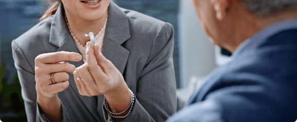 A professional holding a small blue Yes Hearing hearing aid while explaining its features to an older man during a consultation. The image emphasizes personalized support and accessibility in the hearing aid.