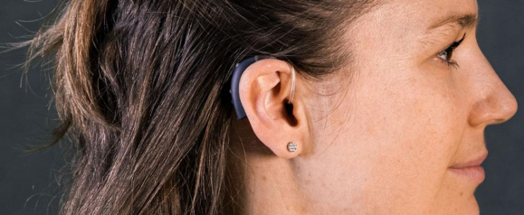 Close-up side profile of a woman wearing a behind-the-ear Beltone hearing aid with a clear tube leading into the ear canal. Her dark hair is pulled back, revealing the device and a small stud earring.