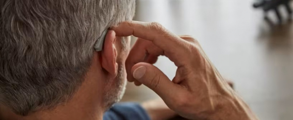 Close-up of an older man adjusting a Bose hearing aid behind his ear with his index finger. The hearing aid is sleek and blends with his gray hair, highlighting its discreet design.