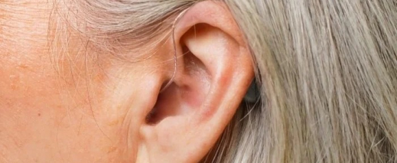 Close-up of a person's ear with a nearly invisible Ceretone hearing aid tucked behind the ear, connected by a thin, clear tube. The image highlights the discreet design of the device, blending seamlessly with the user's light gray hair.