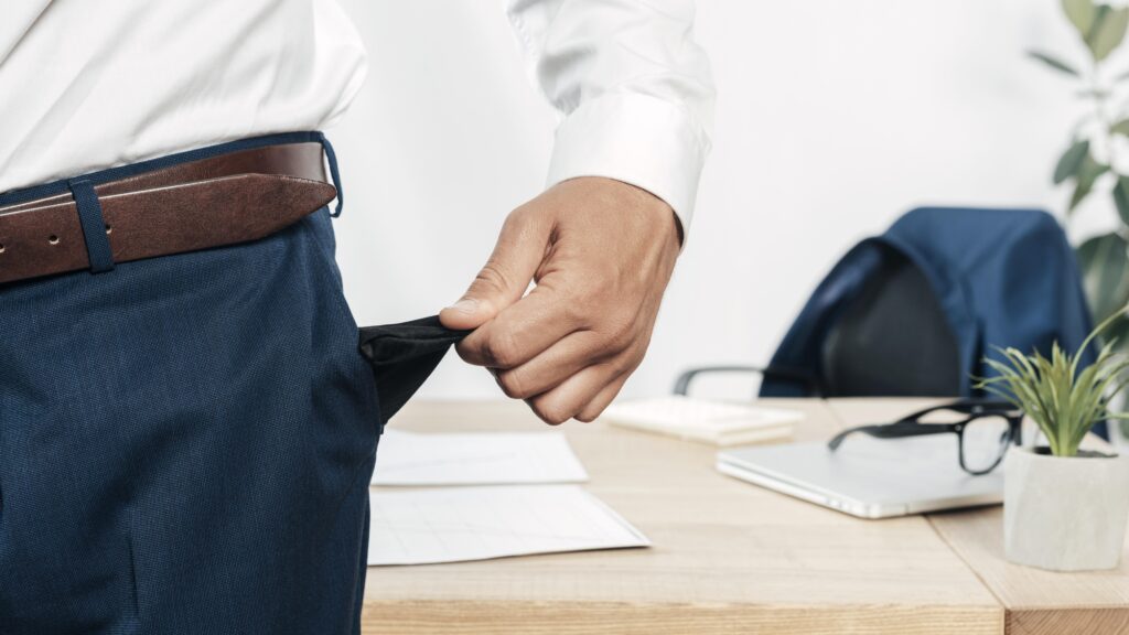 A man in business attire pulling out an empty pocket while standing beside a desk, symbolizing financial distress and illustrating what happens when you file for bankruptcy. The desk holds documents, glasses, a laptop, and a small plant, suggesting a professional setting.