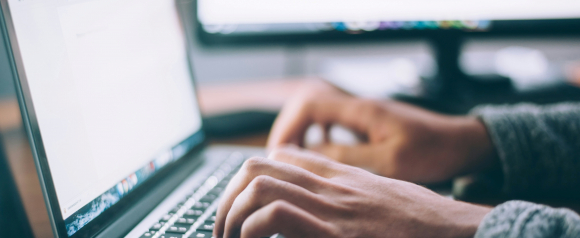 Close-up of a person typing on a laptop keyboard with a blurred monitor in the background. The screen shows a blank or minimal interface, suggesting data entry or software use, possibly within a CRM like Salesforce.