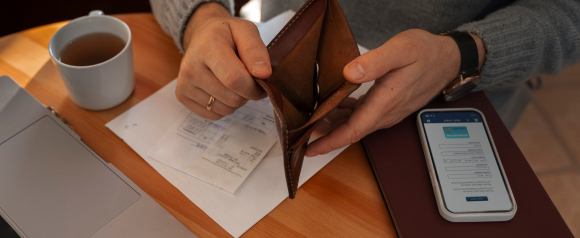 Man holding an empty wallet while sitting at a desk with bills, a cup of tea, a laptop, and a smartphone displaying a Century debt relief form. The scene suggests financial stress and the need for assistance with debt management.