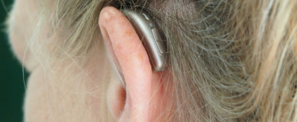 Close-up view of a woman's ear with an OmniHear hearing aid resting behind it. The device is metallic and streamlined, blending subtly with her blonde hair.