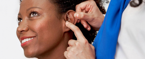 A smiling woman is being fitted with a behind-the-ear hearing aid by a professional wearing a blue and white uniform. The close-up image shows the device being carefully placed on her ear, emphasizing personalized hearing care at HearingLife.