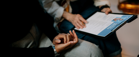 A job interview scene shows one person holding a clipboard with a resume while another sits nearby with hands folded, suggesting an HR discussion or candidate review. The image supports content about HR software like BambooHR by visually representing hiring interviews employee records and human resources workflows.