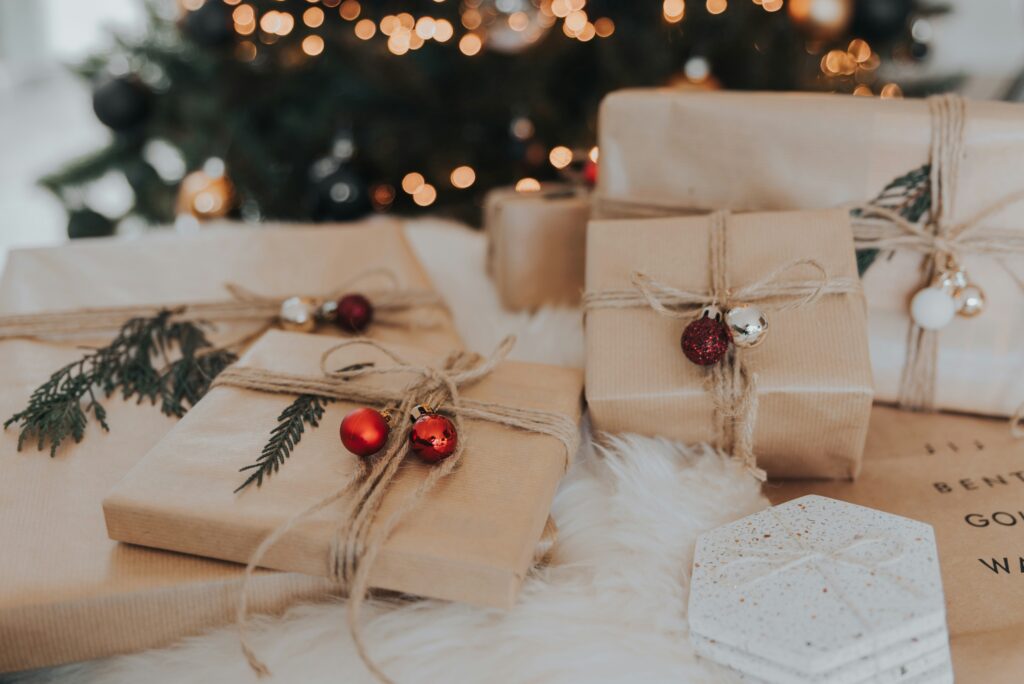 Rustic Christmas gifts wrapped in brown paper with twine and red ornaments under a Christmas tree with bokeh lights.