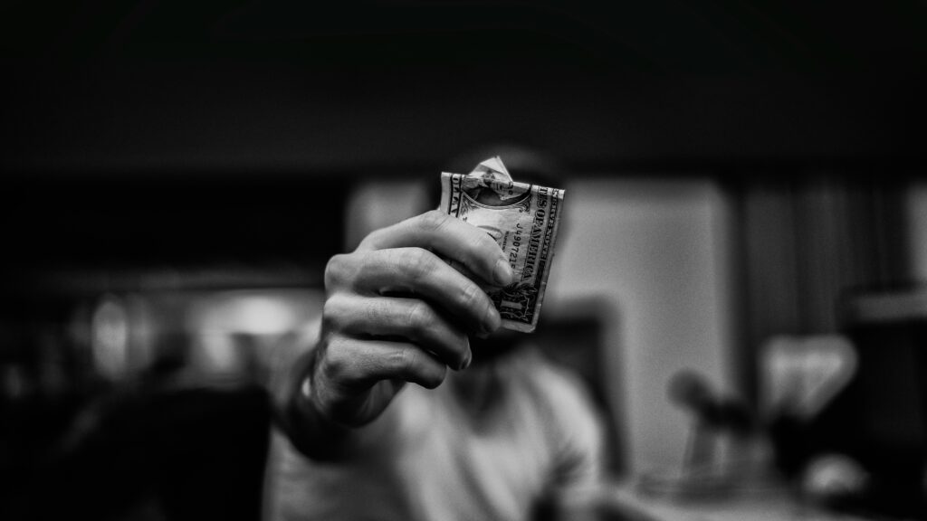 Black and white photo of a person holding out a folded one-dollar bill toward the camera, with the background blurred. The close-up shot emphasizes the concept of money and financial exchange, relevant to discussions about tax relief or the Lifetime Gift Tax Exemption.