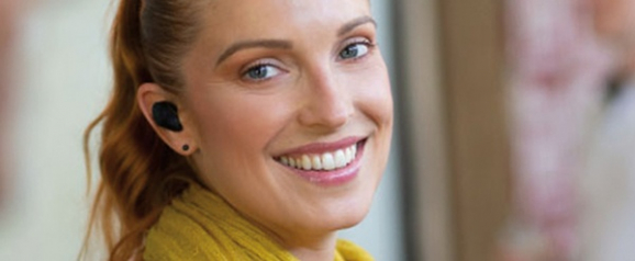 Smiling woman wearing a black Nuheara hearing aid in her ear, with her hair tied back and a yellow scarf around her neck. The image highlights the modern, earbud-style design of the hearing device in a casual, everyday setting.
