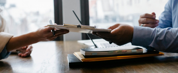 Two people exchange a clipboard with documents across a wooden desk, with notebooks, folders, and a laptop in the background. The setting appears to be a professional office, suggesting a business meeting or onboarding process related to payroll or HR management using ADP payroll software.