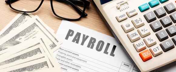 Stacked US dollar bills sit beside a calculator glasses and a printed document labeled PAYROLL on a wooden desk illustrating payroll processing and financial administration. The image visually supports content about payroll software like OnPay by showing tools and paperwork commonly used to calculate and manage employee pay.