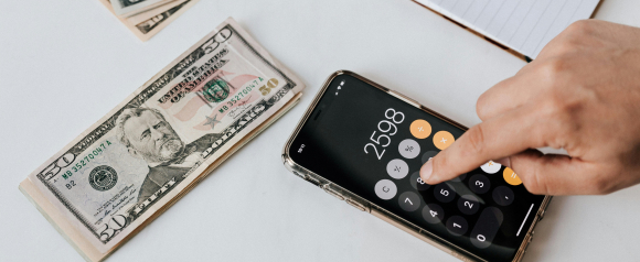 A hand taps numbers into a smartphone calculator next to a stack of US dollar bills on a white desk, suggesting pay calculation or wage verification. The image supports content about payroll software like Paychex by visually representing digital pay calculations and employee compensation management.