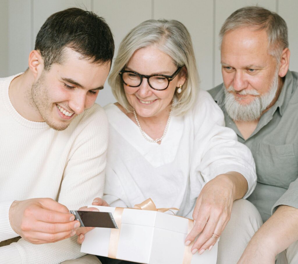 A smiling older couple and a young man sit closely together while opening a white gift box tied with a peach-colored ribbon. The woman, wearing glasses and a pearl necklace, helps hold the box as the young man pulls out a photo or card, suggesting a thoughtful, sentimental present.