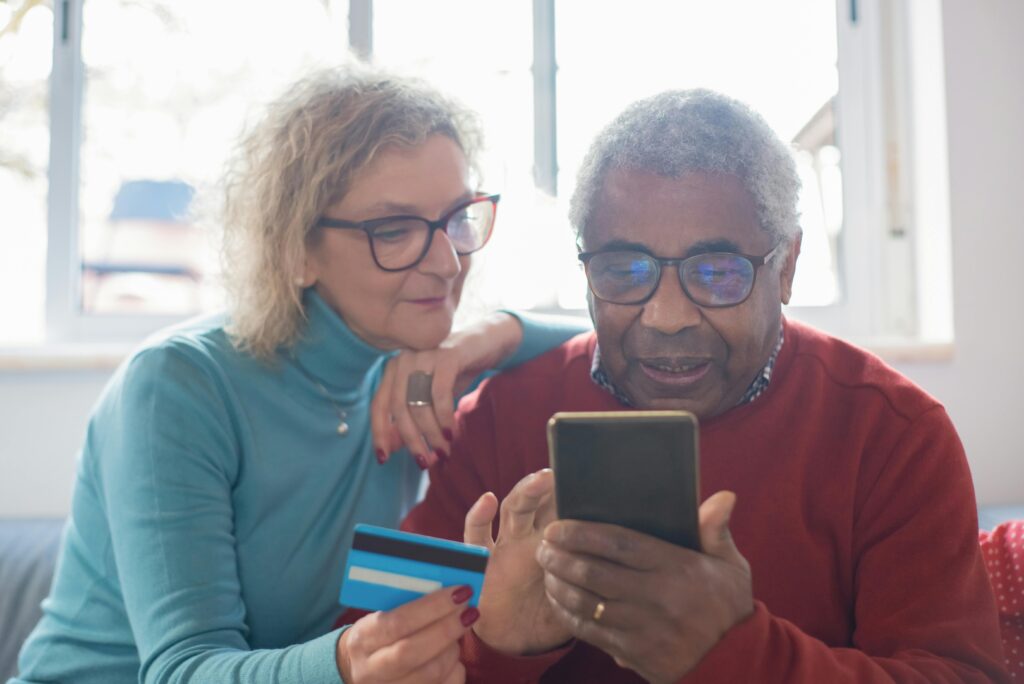 Older couple sitting together on a couch, with the man holding a smartphone and the woman holding a blue-striped credit card, both looking at the screen. They appear focused and engaged, possibly reviewing finances or negotiating credit card debt settlement online.