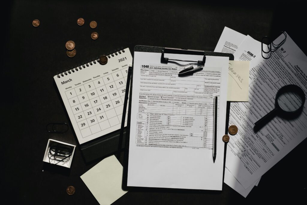 A 1040 tax form sits on a clipboard surrounded by scattered coins, IRS documents, a magnifying glass, and a March 2021 calendar. The scene suggests preparing taxes or responding to a letter from the IRS, tying into themes of tax relief.