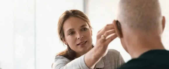 Woman gently adjusting a Sennheiser hearing aid on an older man's ear in a well-lit room. The image captures a moment of care and professional support, emphasizing the user-friendly design of the hearing device.