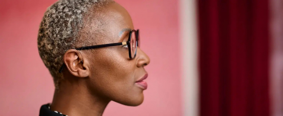 Side profile of a woman wearing glasses and a discreet Signia hearing aid behind her ear, against a pink and burgundy background. The image highlights the sleek, modern design of hearing AI technology in a lifestyle setting.