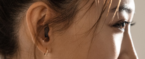 Close-up of a woman’s ear wearing a discreet, dark-colored Sony hearing aid, with part of her smiling face and eye in soft focus. The image highlights the subtle design and comfortable fit of modern hearing aid technology.