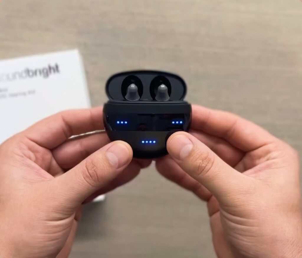 Hands holding the open charging case of Soundbright Mini hearing aids displaying blue LED battery indicators, illustrating the hardware design for Soundbright Mini Reviews.