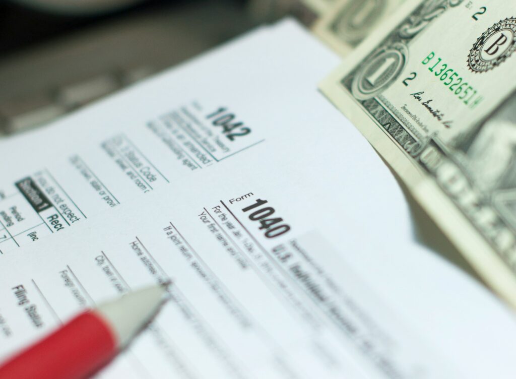 Alt text: Close-up of U.S. tax forms 1040 and 1042 partially filled out, with a red pen pointing toward one section and a few dollar bills visible in the corner. The image suggests preparation for the tax deadline.