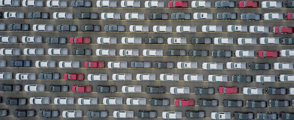 Aerial view of a large parking lot filled with rows of parked pickup trucks in mostly white, gray, and black, with a few red vehicles standing out in a pattern. The image conveys vehicle organization and inventory scale, suitable for themes like fleet management or vehicle tracking.