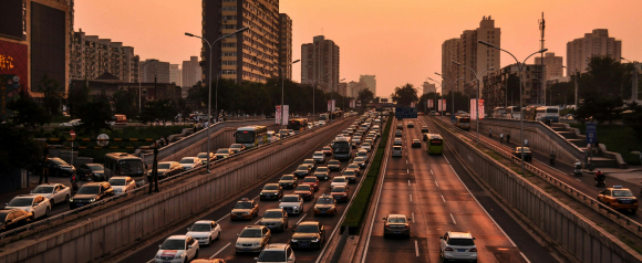 Dense city traffic fills multiple lanes on a divided highway at sunset, with cars, buses, and taxis moving slowly between tall buildings. Streetlights and signs line the road, capturing the urban congestion and movement typical for vehicle tracking systems like Catrak.