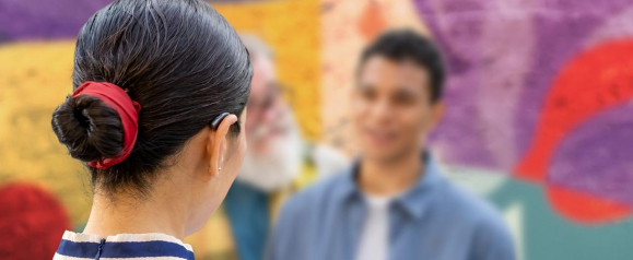 Rear view of a woman with dark hair in a bun secured by a red tie, wearing a black WS Audiology hearing aid while speaking to two blurred individuals in front of a colorful mural. The image illustrates social interaction supported by advanced hearing technology in a vibrant, everyday setting.