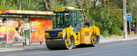 A yellow XCMG road roller compacts fresh asphalt near a bus stop with bright Fanta advertisements, surrounded by trees and parked cars. The image highlights how FleetWatcher fleet tracking helps monitor and optimize heavy equipment usage in urban infrastructure projects.