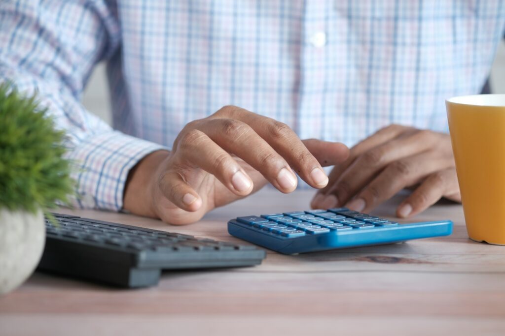Close-up of hands using a blue calculator on a wooden desk to estimate total tax liabilities and federal income tax due.
