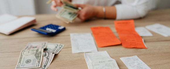 Person organizing paper receipts and U.S. dollar bills on a wooden table, with a blue calculator and open notebook nearby. The scene illustrates hands-on budgeting or expense tracking, supporting themes of debt relief and financial planning with services like InCharge Debt Solutions.
