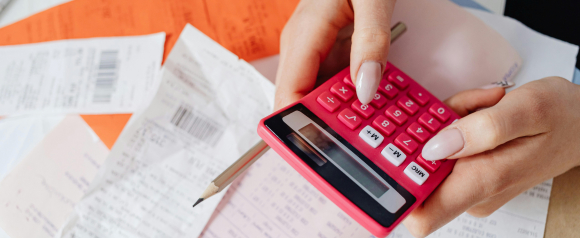 Close-up of hands using a pink calculator with a pencil, surrounded by various receipts and financial documents. The image highlights personal expense tracking and budgeting, aligning with debt relief solutions offered by companies like PDS Debt.